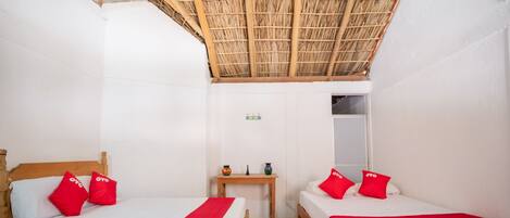 A room with two beds, each with red pillows and a white bedspread. There is a small wooden table between the beds with a vase and a candle. The room has a thatched roof ceiling and white walls.