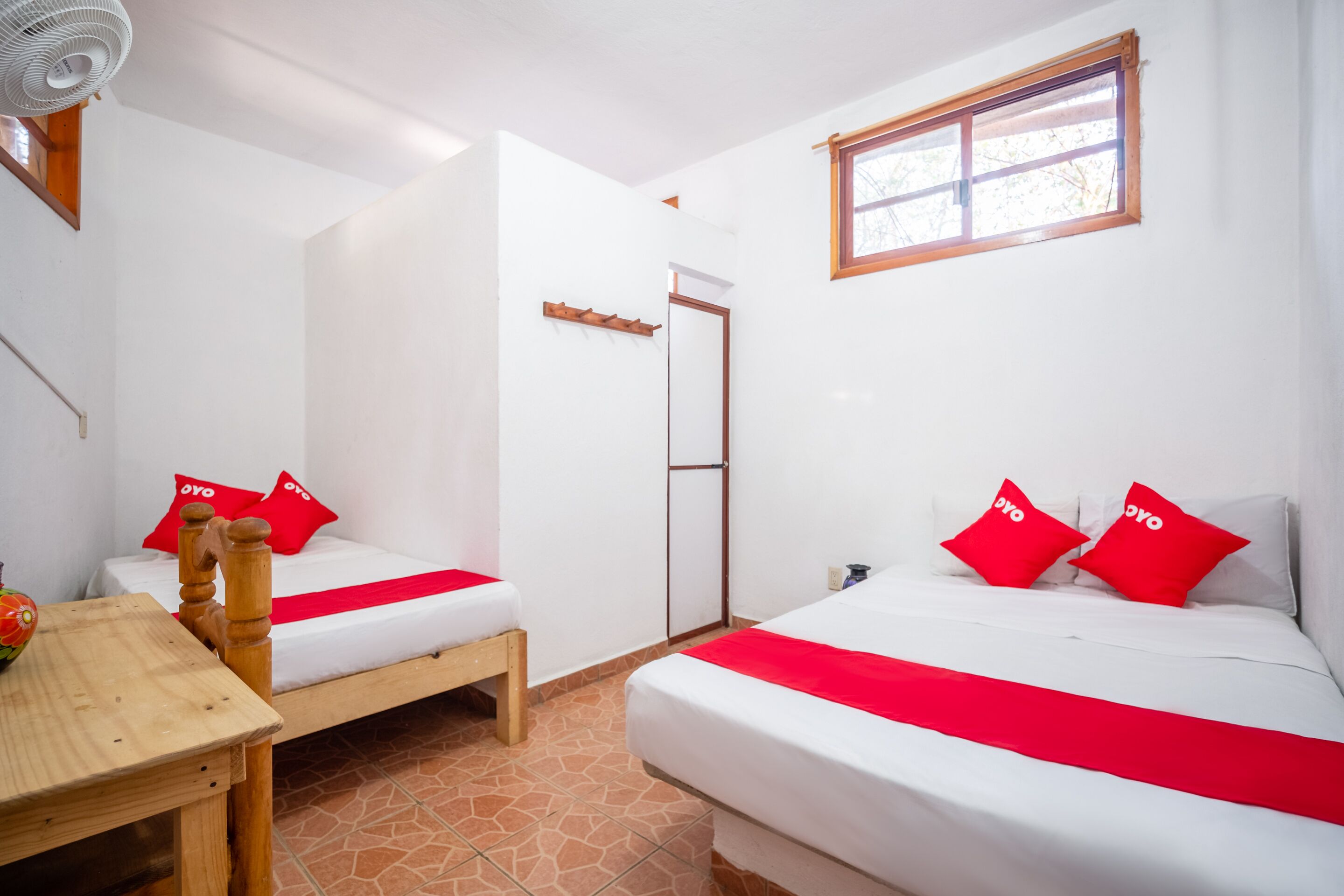 Camera d'albergo con due letti singoli, ciascuno con copripiumino e cuscini rossi. Si nota un tavolo in legno e una sedia, una finestra con cornice in legno e una ventola a soffitto.