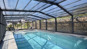 A covered swimming pool with clear glass panels, stone walls, and lounge chairs. - Luxurious Villa in Nadrin Belgium with Sauna & Hot Tub (Houffalize)