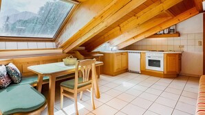 Uma cozinha acolhedora com vigas de madeira, uma mesa de jantar, cadeiras, forno embutido e uma janela com vista para uma floresta.