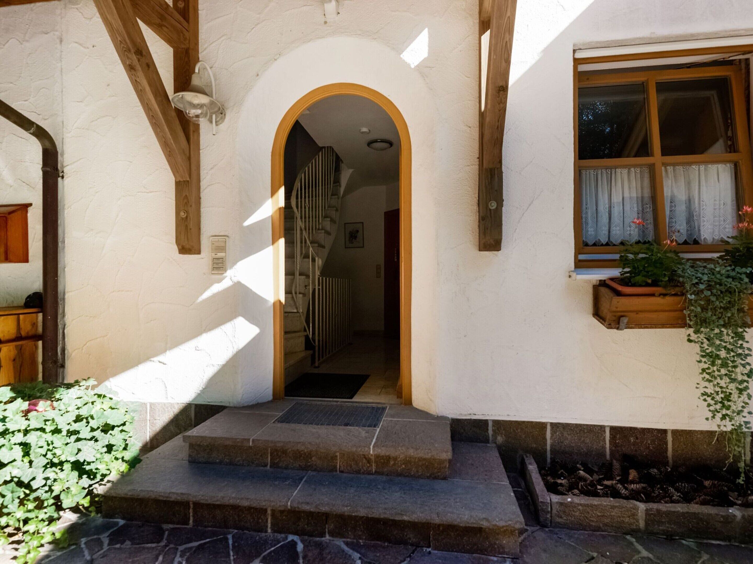 Una escalera de piedra que conduce a una entrada arqueada, una ventana de madera con cortinas y una planta en maceta a la derecha.