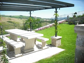 Un patio con una gran mesa y bancos, rodeado por un muro de piedra y vistas a colinas onduladas.