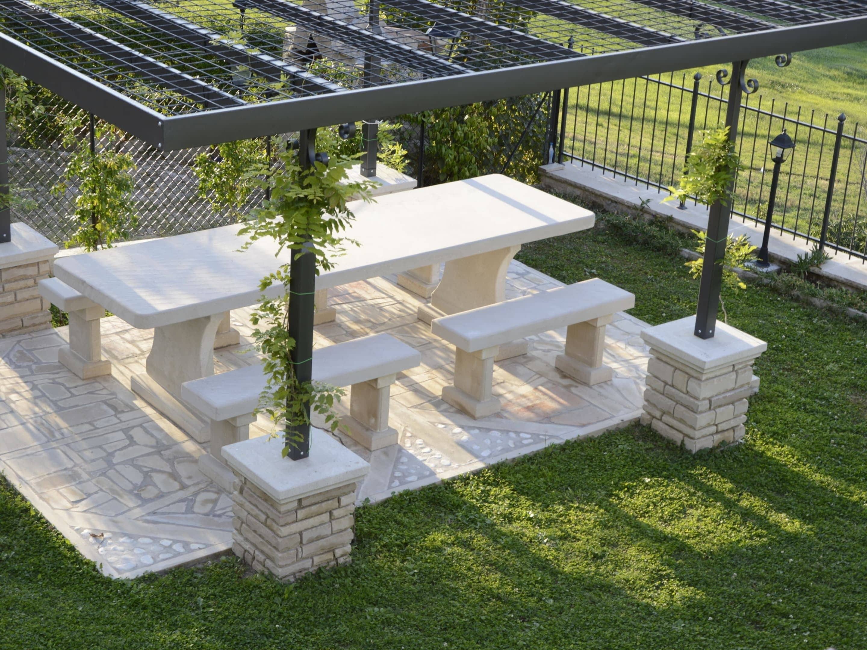 An outdoor patio with a stone table and benches, surrounded by a metal fence and greenery.