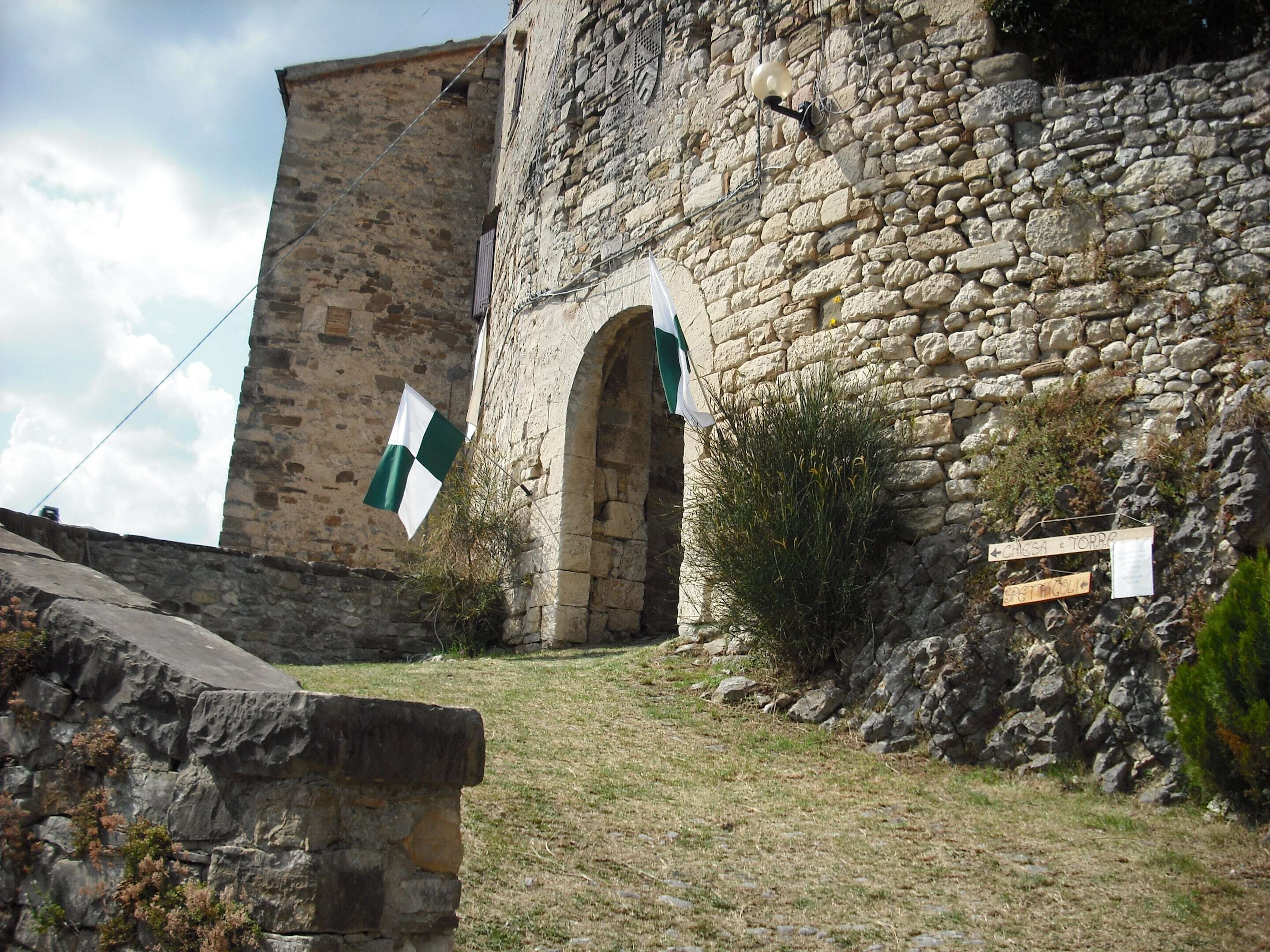 Un edificio de piedra con una torre, una bandera y un cartel con indicaciones.