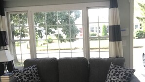 Ein Wohnzimmer mit grauem Ecksofa, zwei gemusterten Zierkissen, einem schwarzen Couchtisch mit einem gewebten Korb und einem Fenster mit Blick auf eine Straße und Bäume.