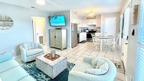 Ein Wohnzimmer mit einer Couch, Sesseln, einem Couchtisch und einem Fernseher. Es gibt eine Küchenzeile mit Schränken, einem Kühlschrank und einem Esstisch mit Stühlen.