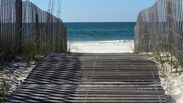 Ein Holzsteg führt zu einem Sandstrand bei blauem Himmel.