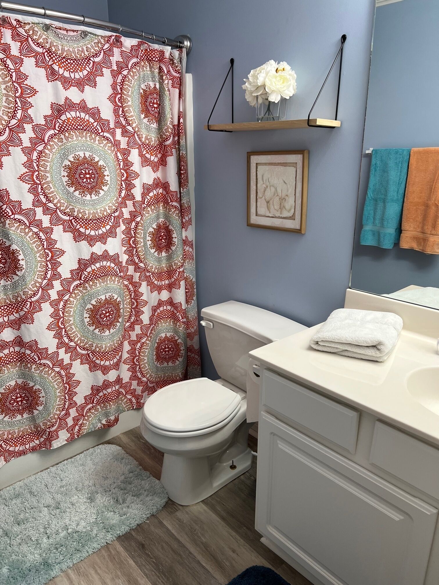 Une salle de bain avec un rideau de douche à motifs, des toilettes, un lavabo avec une serviette et une étagère sur laquelle se trouvent un vase et un tableau encadré.