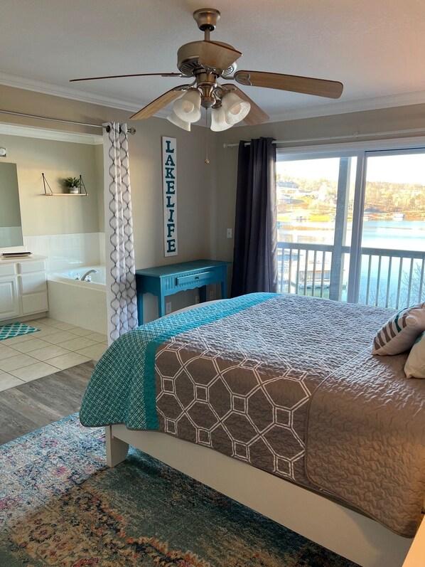 A bedroom with a bed, a ceiling fan, a window with a view of a lake, and a wall-mounted shelf with a plant.