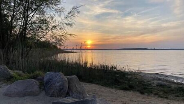 Un coucher de soleil sur une étendue d’eau calme, bordée d’une côte rocheuse et d’une végétation clairsemée.