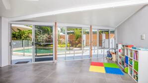 A spacious room with a large sliding glass door leading to a pool area, a ceiling fan, and a children's bookshelf. - Tropical Beachside Home - Pool, Wifi (Holloways Beach)