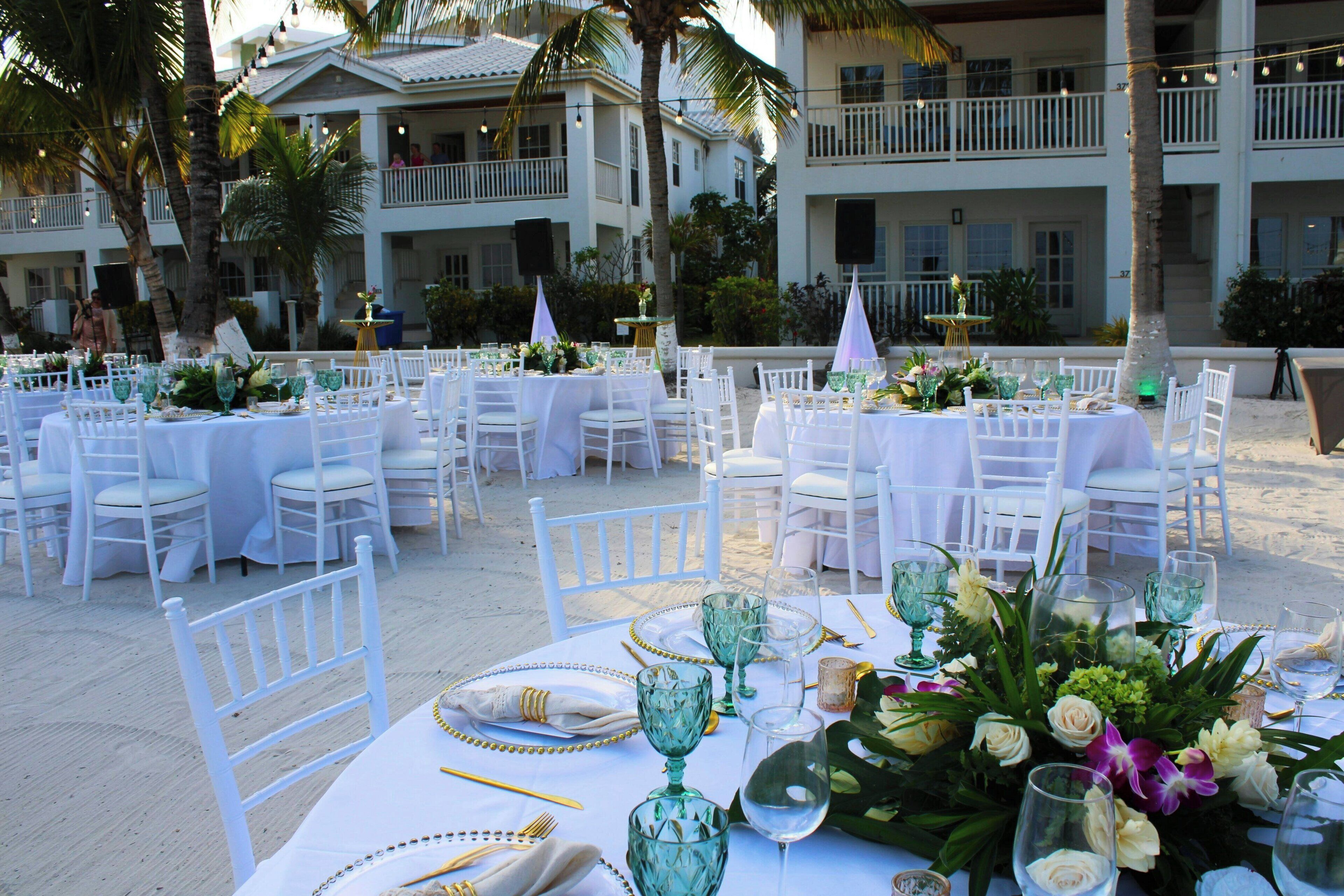 An outdoor dining area with white tables and chairs, surrounded by palm trees and a building with balconies.