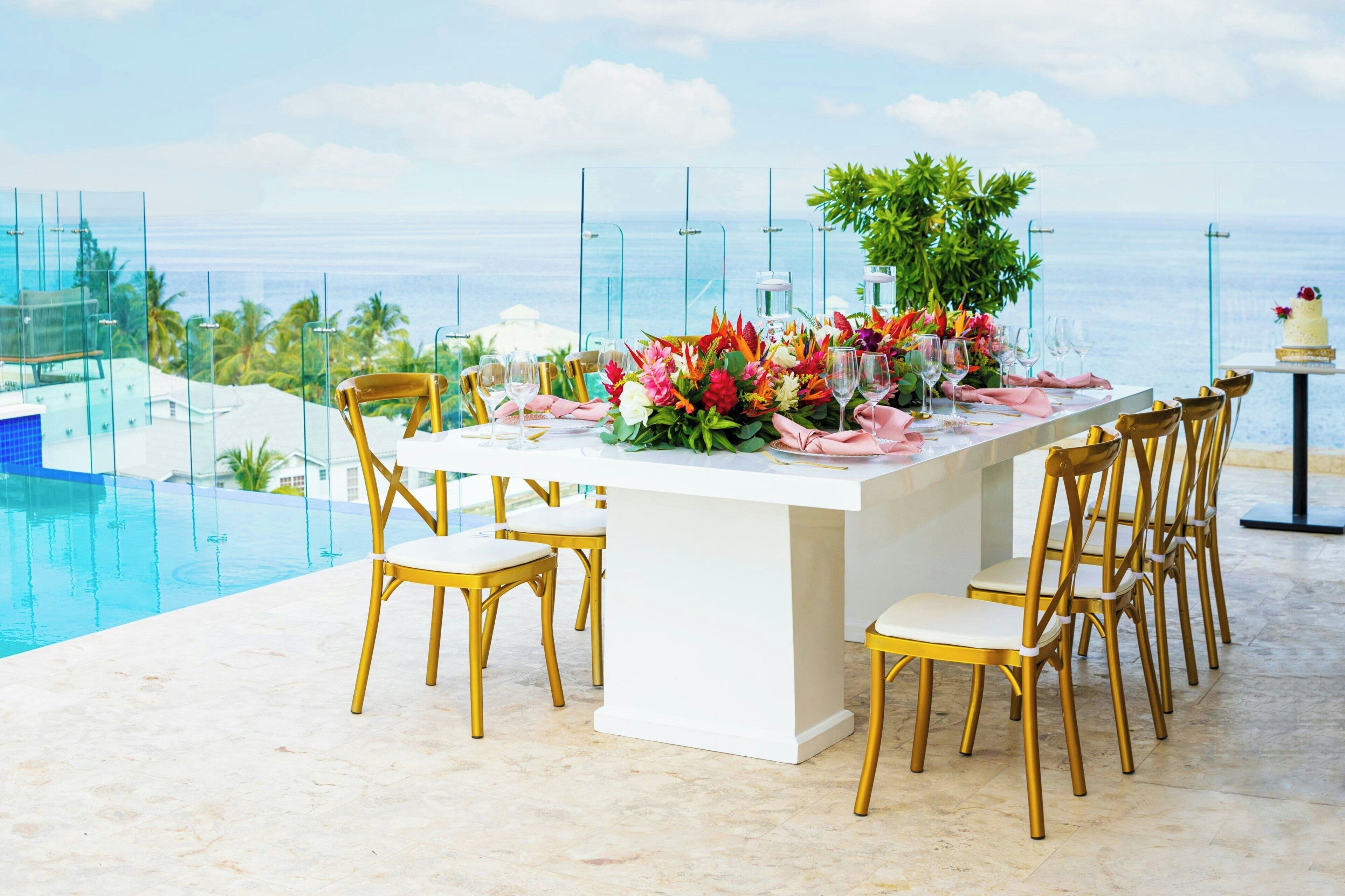 A dining area with a white table set for six, surrounded by golden chairs, overlooking a pool and ocean view.