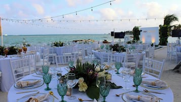 An outdoor wedding setup with round tables, white chairs, and string lights. Tables are set with glassware, plates, and napkins.