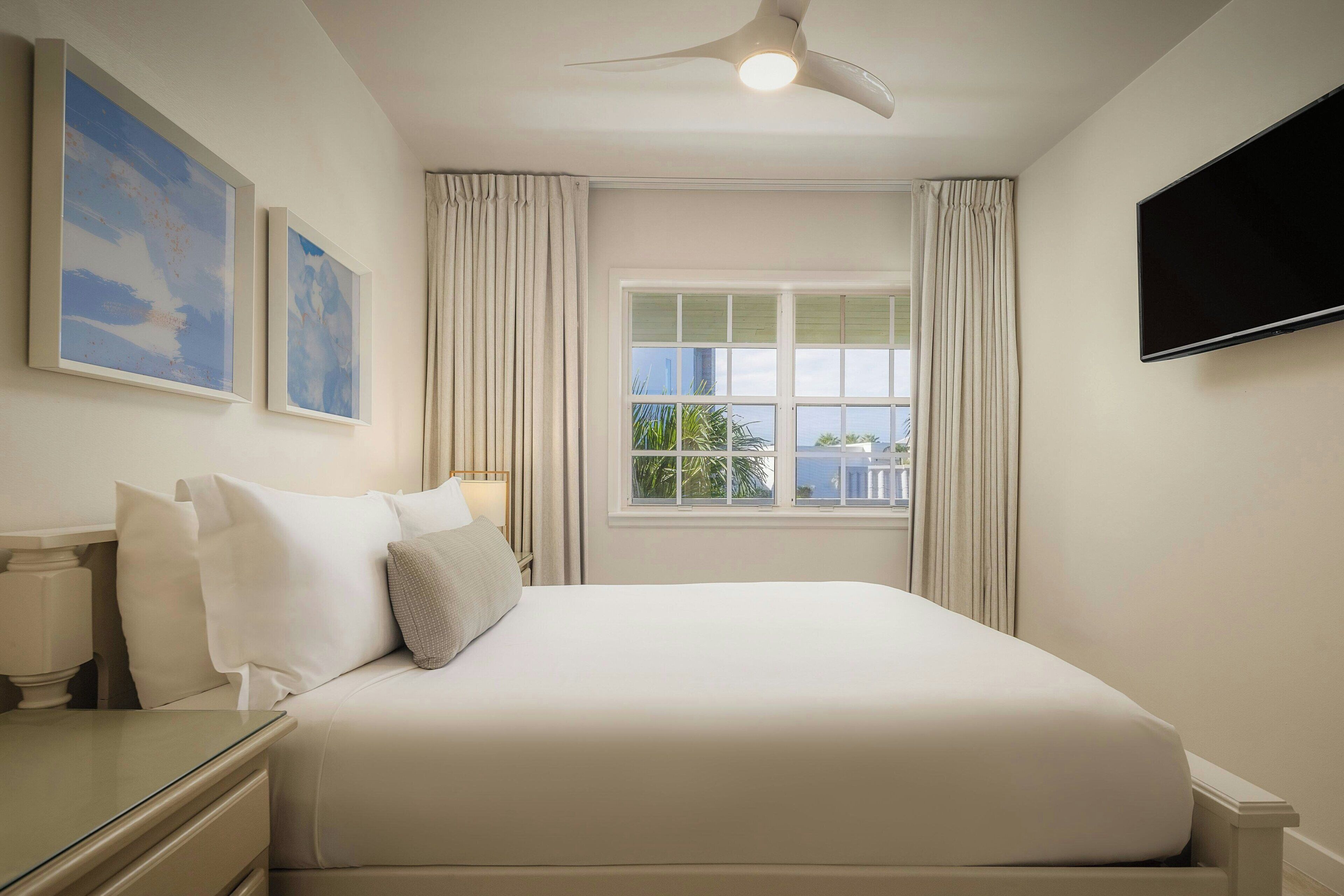 Habitación de hotel con cama, mesita de noche, lámpara, televisor de pantalla plana en la pared y ventana con cortinas.