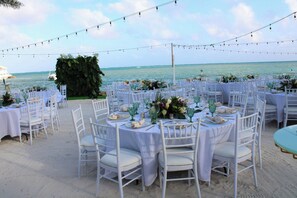 An outdoor dining area with round tables set for a meal, surrounded by white chairs, with a view of the ocean and boats in the background.