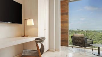 A modern room with a flat-screen TV, a desk, a chair, a lamp, and a view of greenery through a large window.