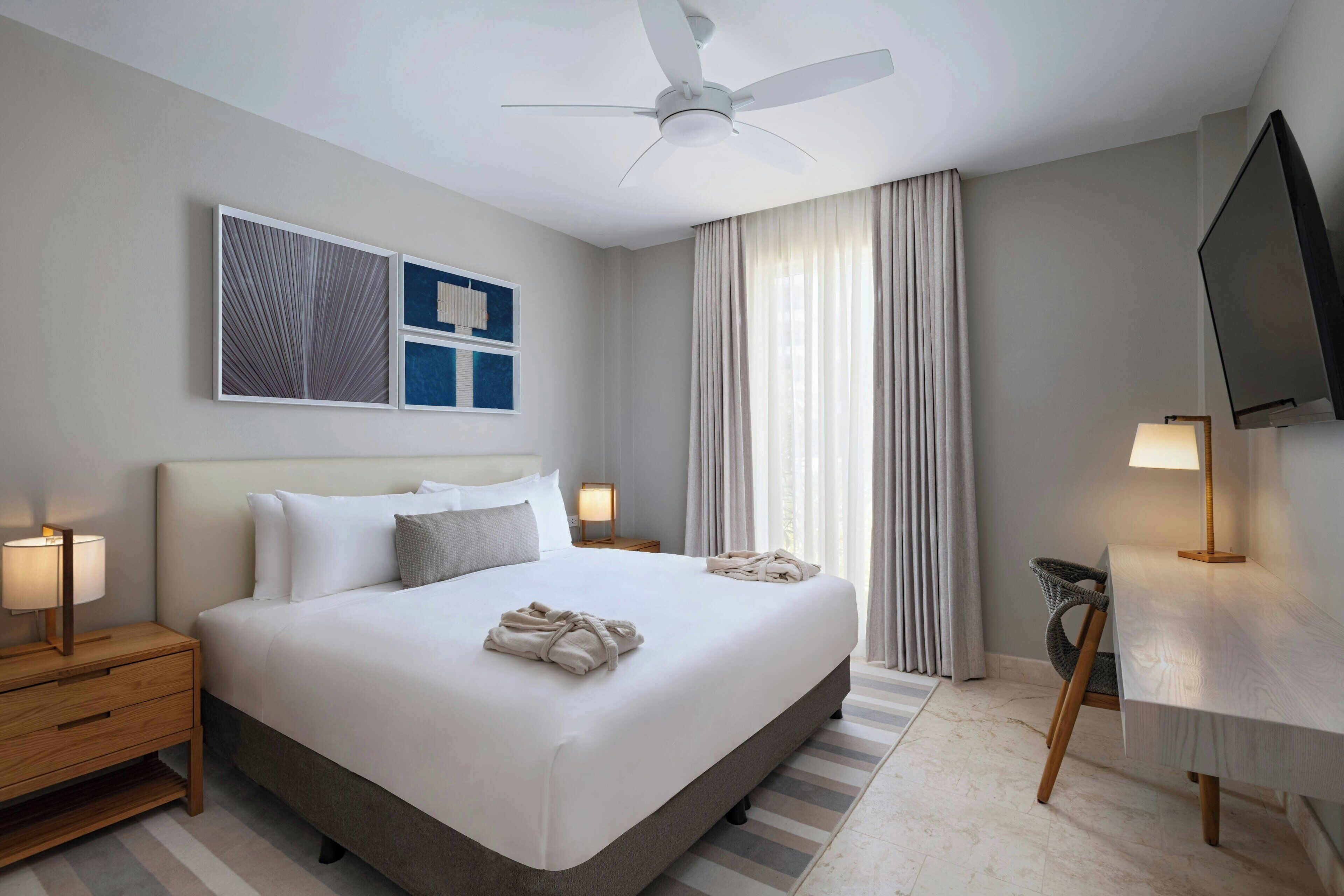 A hotel room with a bed, bedside tables, a ceiling fan, a TV, and artwork on the wall.