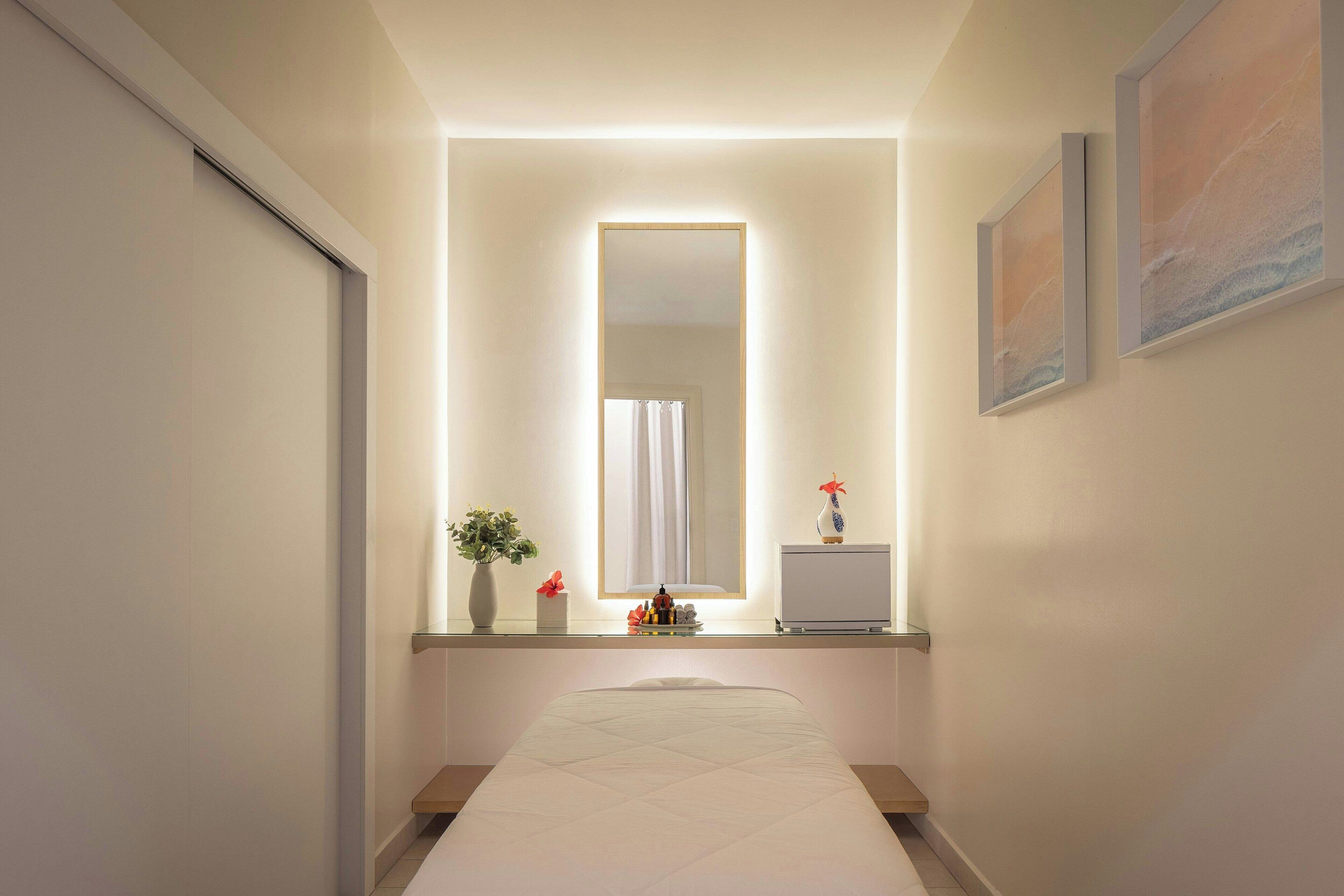 A hallway with a bed, a mirror, a vase with flowers, and a small table with toiletries.