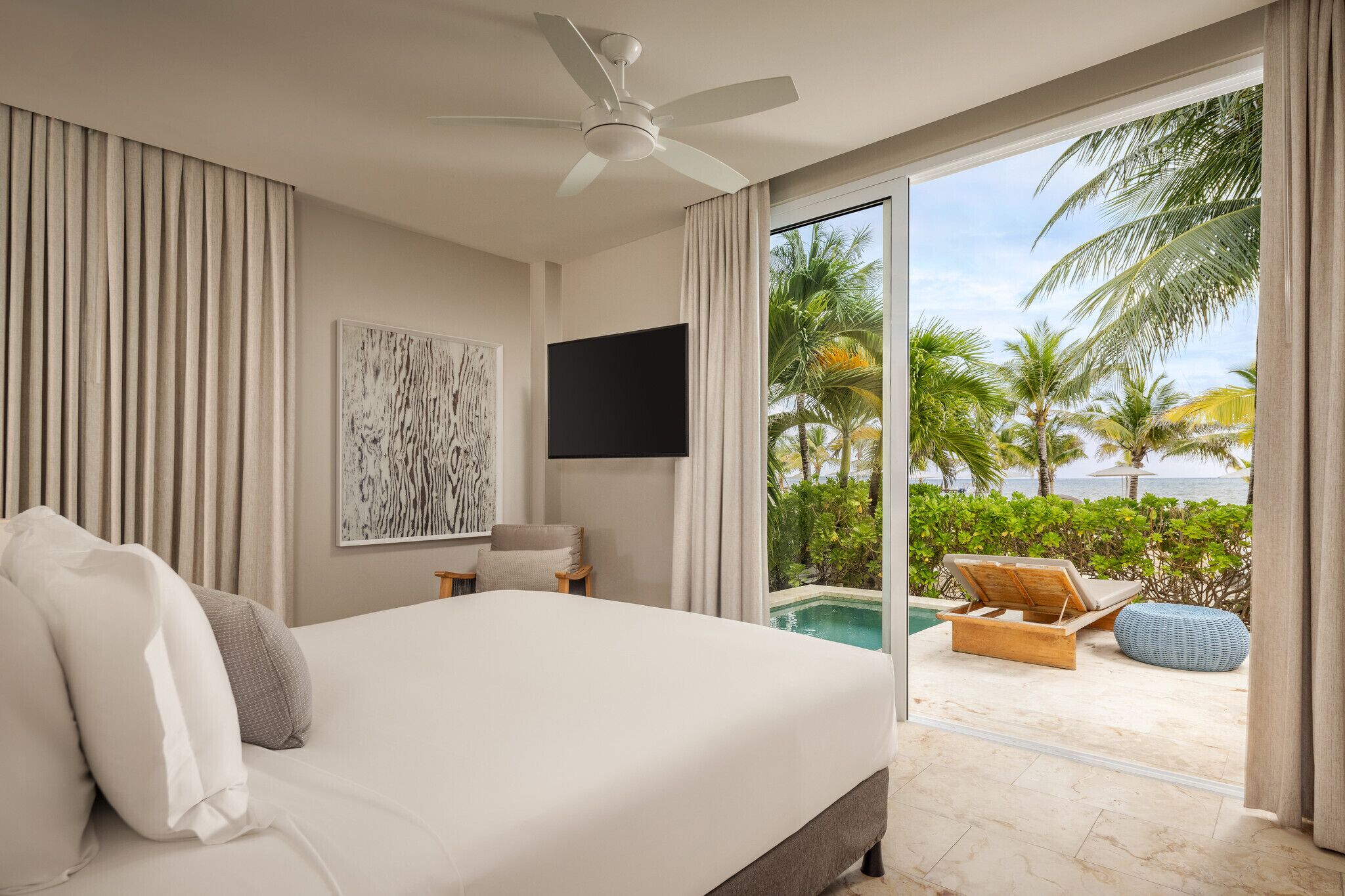 A bedroom with a large bed, a ceiling fan, a flat-screen TV, and a view of a pool and palm trees.
