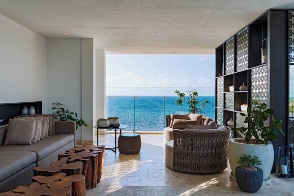 A modern living room with a large window overlooking the sea, a sofa, a round wicker chair, and a bookshelf. - Alaia Belize, Autograph Collection  (San Pedro)