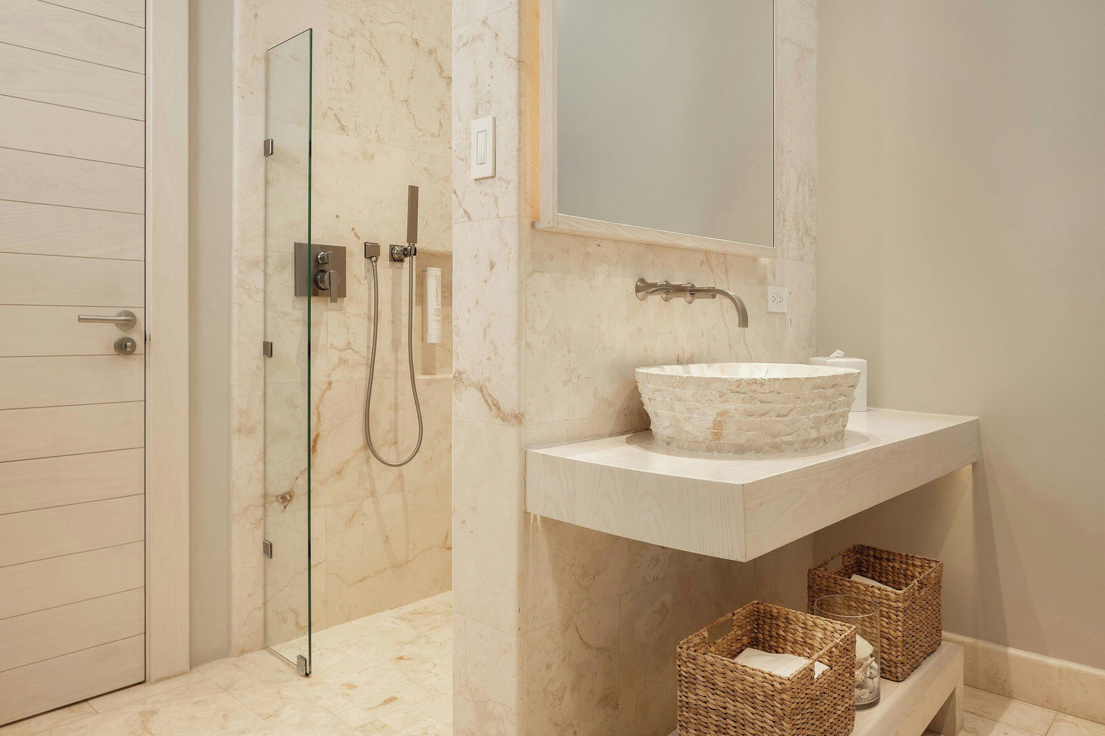 A bathroom with a glass shower enclosure, a marble-tiled floor, a white sink with a stone basin, and wicker baskets on a shelf.