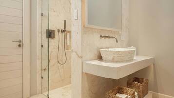 A bathroom with a glass shower enclosure, a marble-tiled floor, a white sink with a stone basin, and wicker baskets on a shelf.