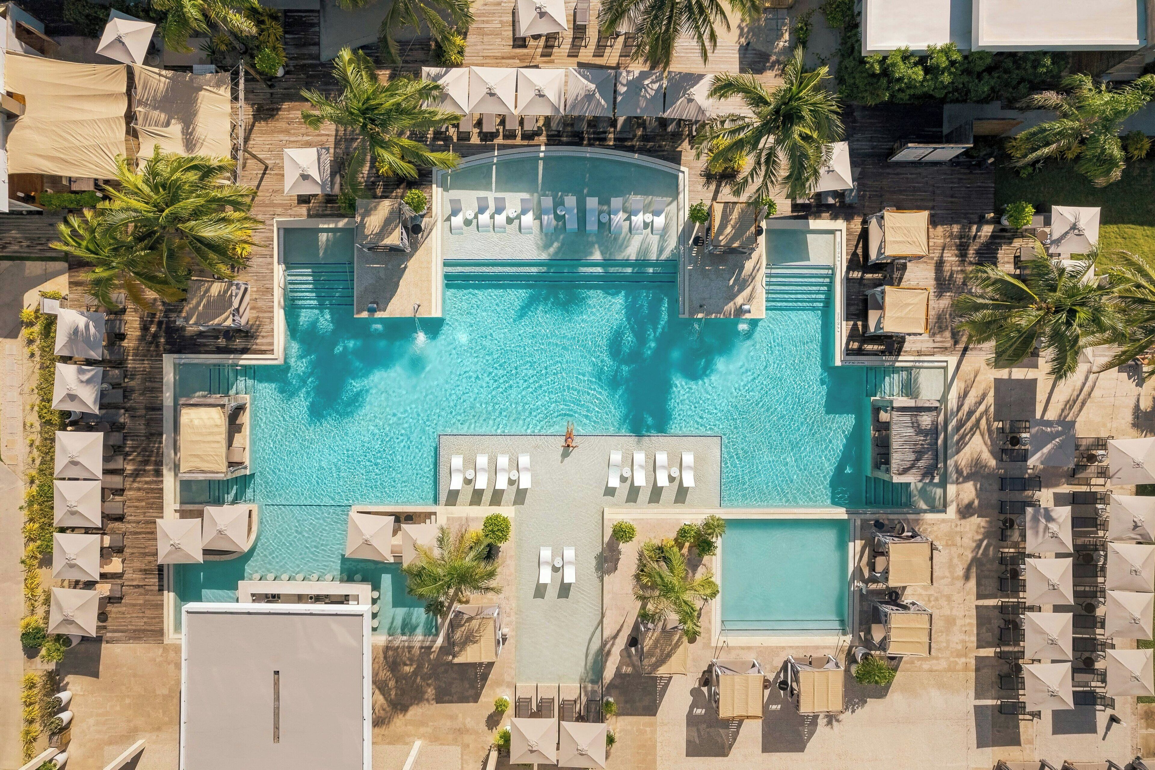 An aerial view of a resort with a central swimming pool, multiple lounge areas, and palm trees.
