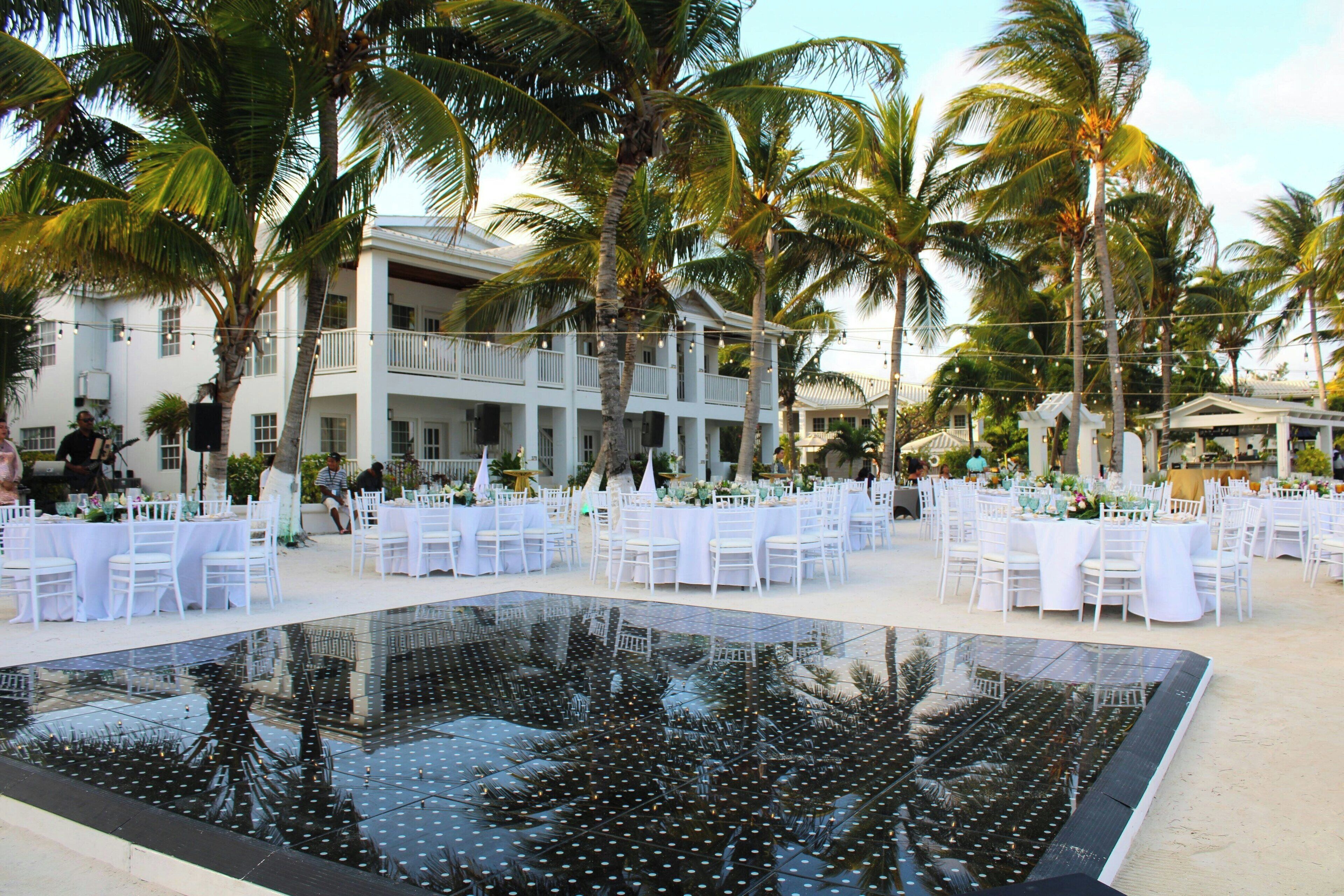 A pool with a glass bottom, surrounded by white tables and chairs, palm trees, and a building with balconies.