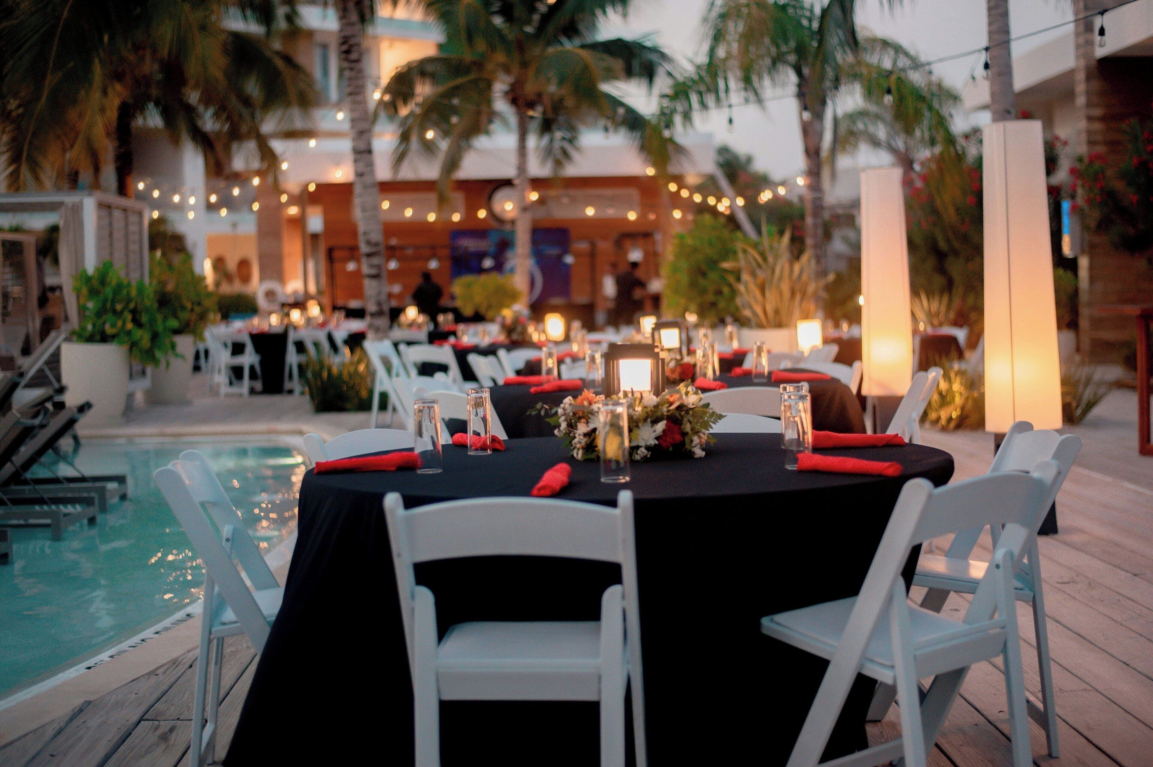An outdoor dining area with tables set for two, surrounded by palm trees and a pool.