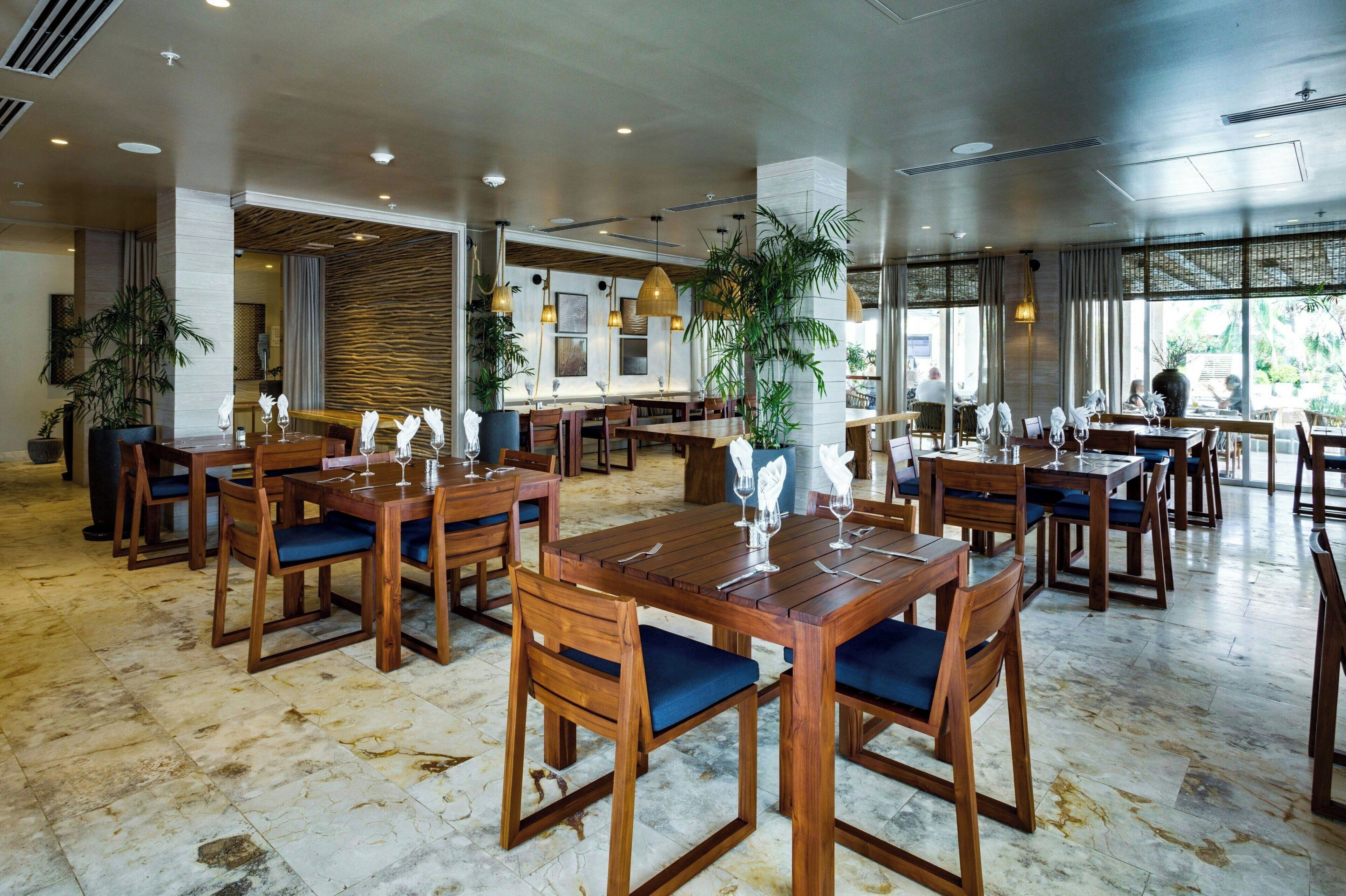 A spacious dining area with wooden tables and chairs, large windows, and indoor plants.