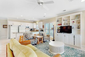 A spacious living room with a sofa, two patterned armchairs, a coffee table, and a television on a stand. The room has white cabinetry and a ceiling fan.