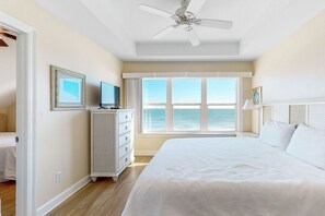 A bedroom with a large bed, a ceiling fan, a dresser, a television, and a window with a view of the ocean.