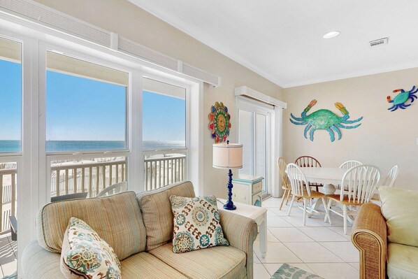 A spacious living room with a large window overlooking the beach, a dining area with white chairs, and a colorful crab wall decoration.