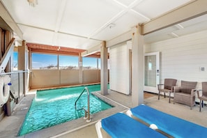 An indoor swimming pool with a view of the beach, a lounge area with chairs, and a door leading to another room.