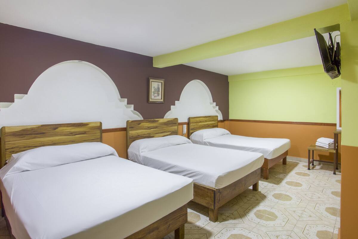 Una habitación de hotel con tres camas de madera, una mesita pequeña y un televisor montado en la pared.