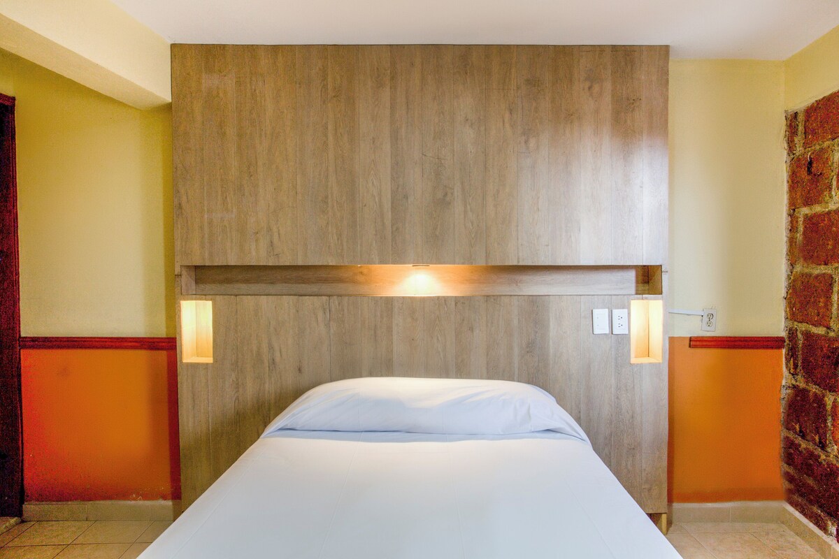 Una habitación de hotel con cabecera de madera, una cama con ropa blanca y dos lámparas fijadas a la pared.