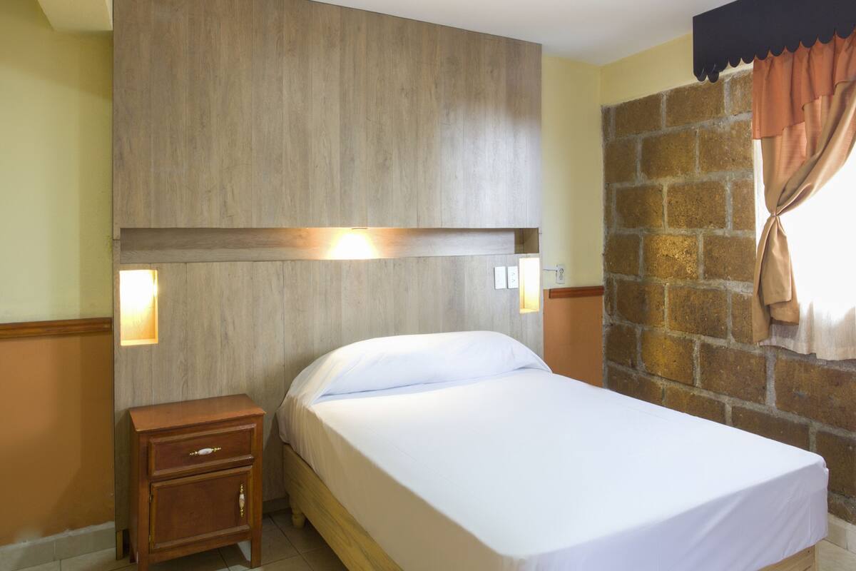 Habitación de hotel con cabecera de madera, una cama individual, una mesita de noche y una pared de ladrillo.