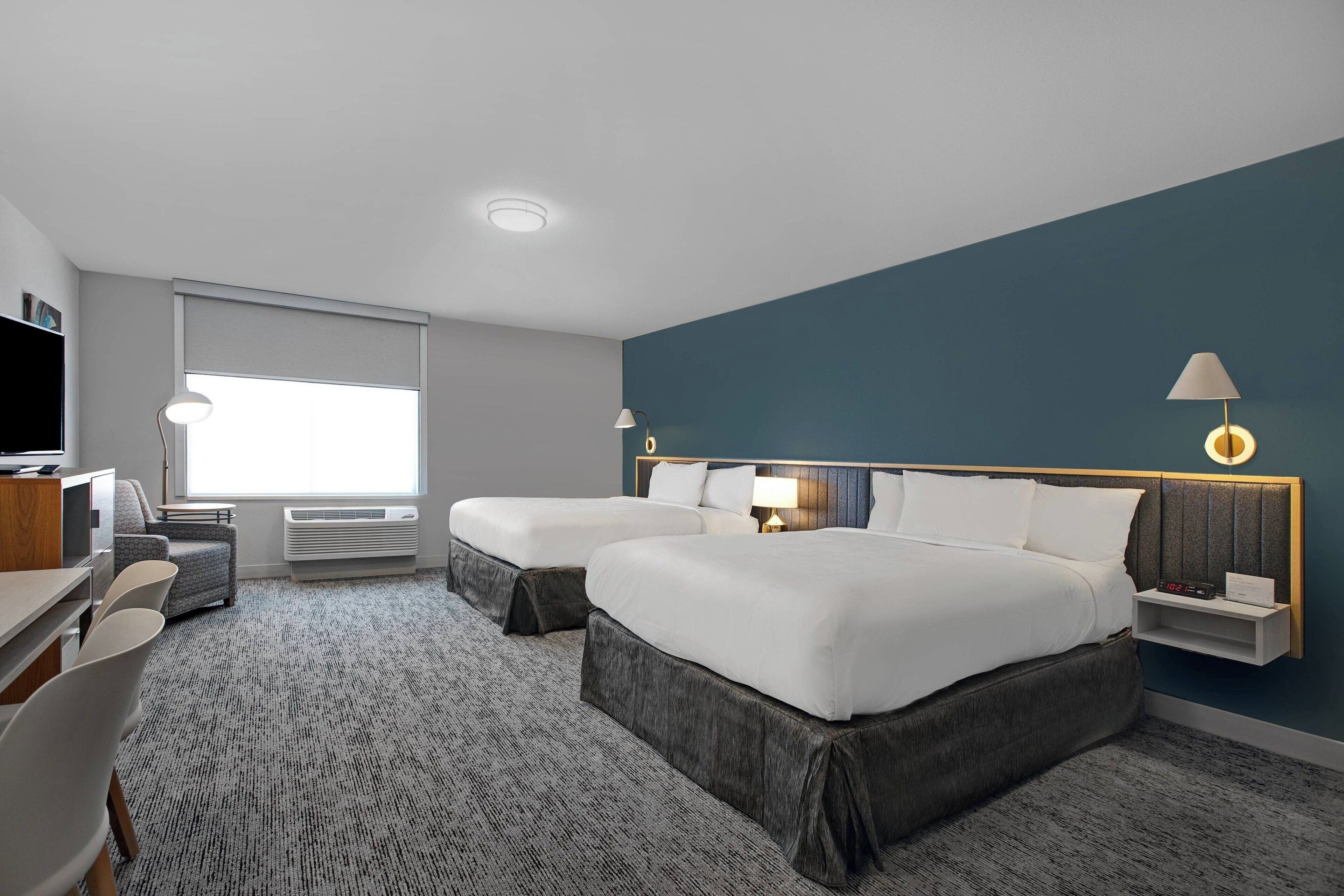 A modern hotel room with two beds, a desk, a chair, and a large window.