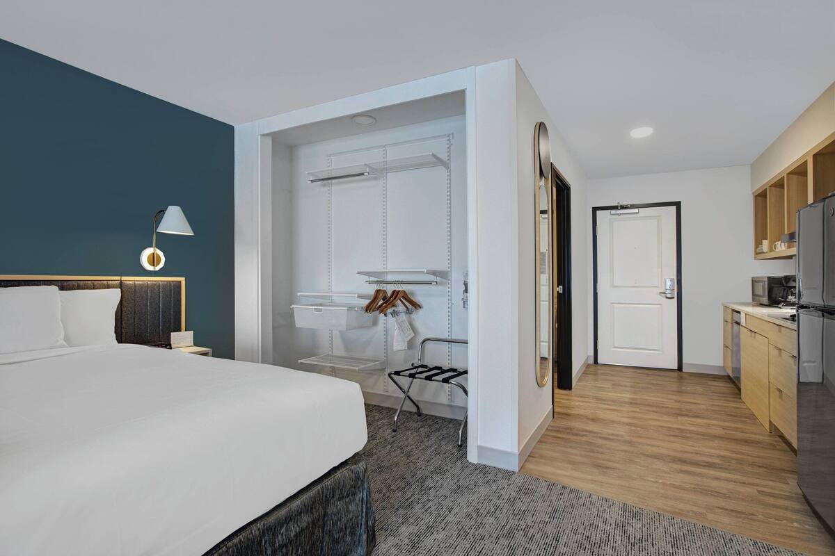 Una habitación de hotel moderna con una cama grande, una lámpara de noche, un armario empotrado, un baño pequeño con espejo y una kitchenette.