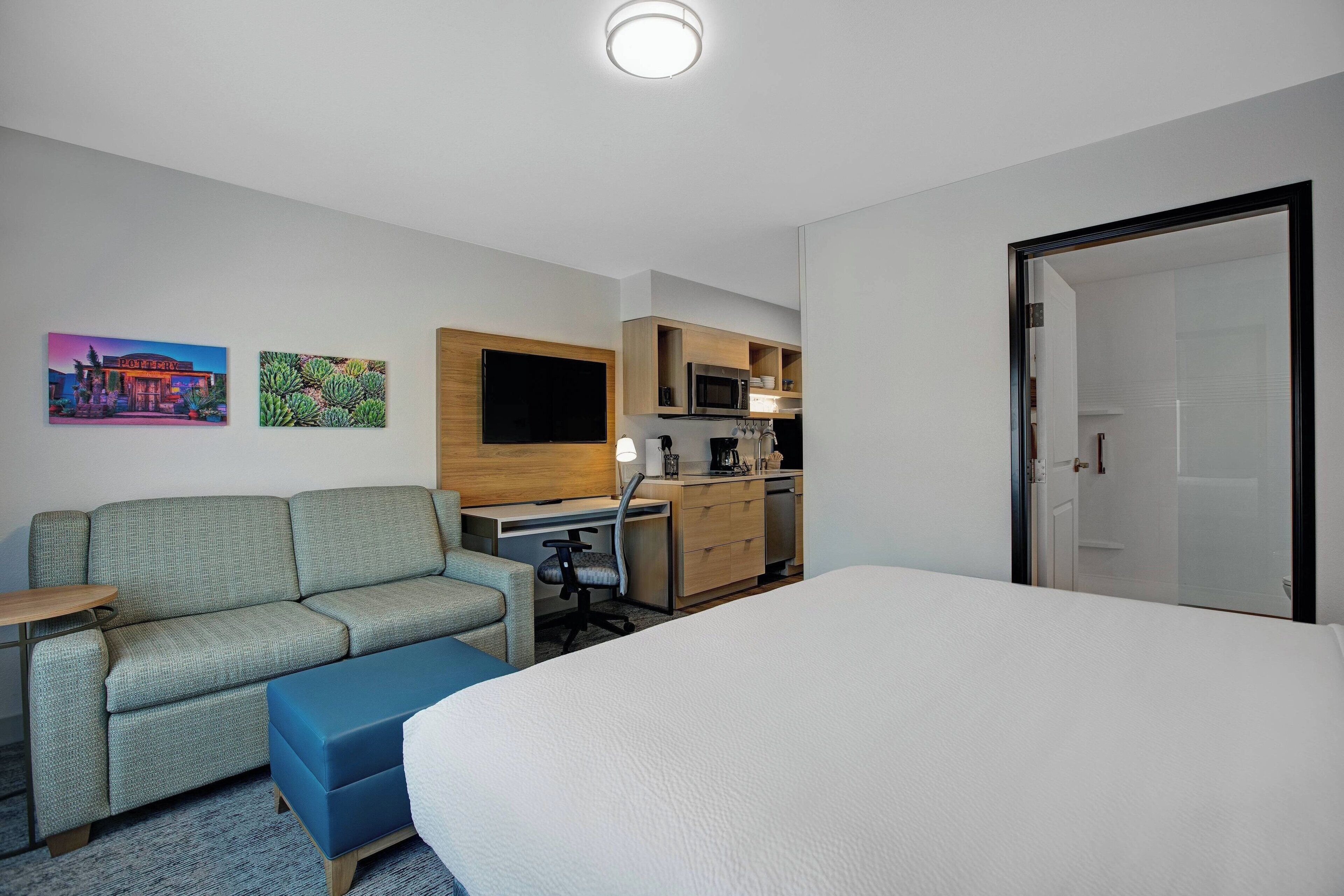 A hotel room with a bed, a sofa, a desk, a chair, a small table, a microwave, a coffee maker, and a door.