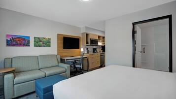 A hotel room with a bed, a sofa, a desk, a chair, a small table, a microwave, a coffee maker, and a door.