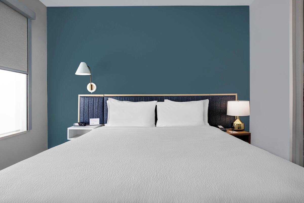 Una habitación de hotel con una cama grande, un cabecero con diseño circular, una mesita de noche con lámpara y una ventana con persianas.