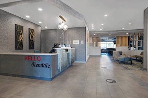 A reception area with a modern design, featuring a reception desk labeled "HELLO Glendale," a seating area with tables and chairs, and a view of the reception desk through a glass partition.