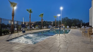 Piscina exterior con jacuzzi, rodeada de sillones de descanso y palmeras, bajo un cielo despejado nocturno.
