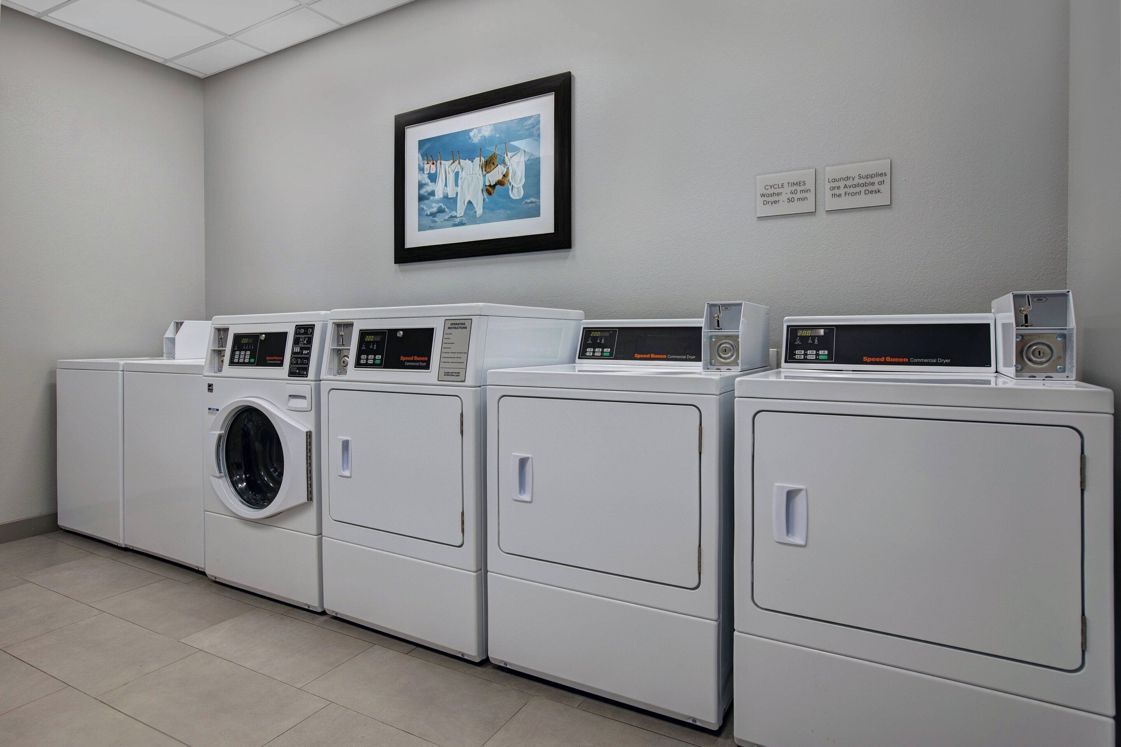 A laundry facility with multiple washing machines and dryers, a framed picture on the wall, and informational signs about cycle times and available supplies.