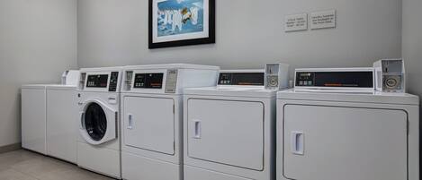 A laundry facility with multiple washing machines and dryers, a framed picture on the wall, and informational signs about cycle times and available supplies.