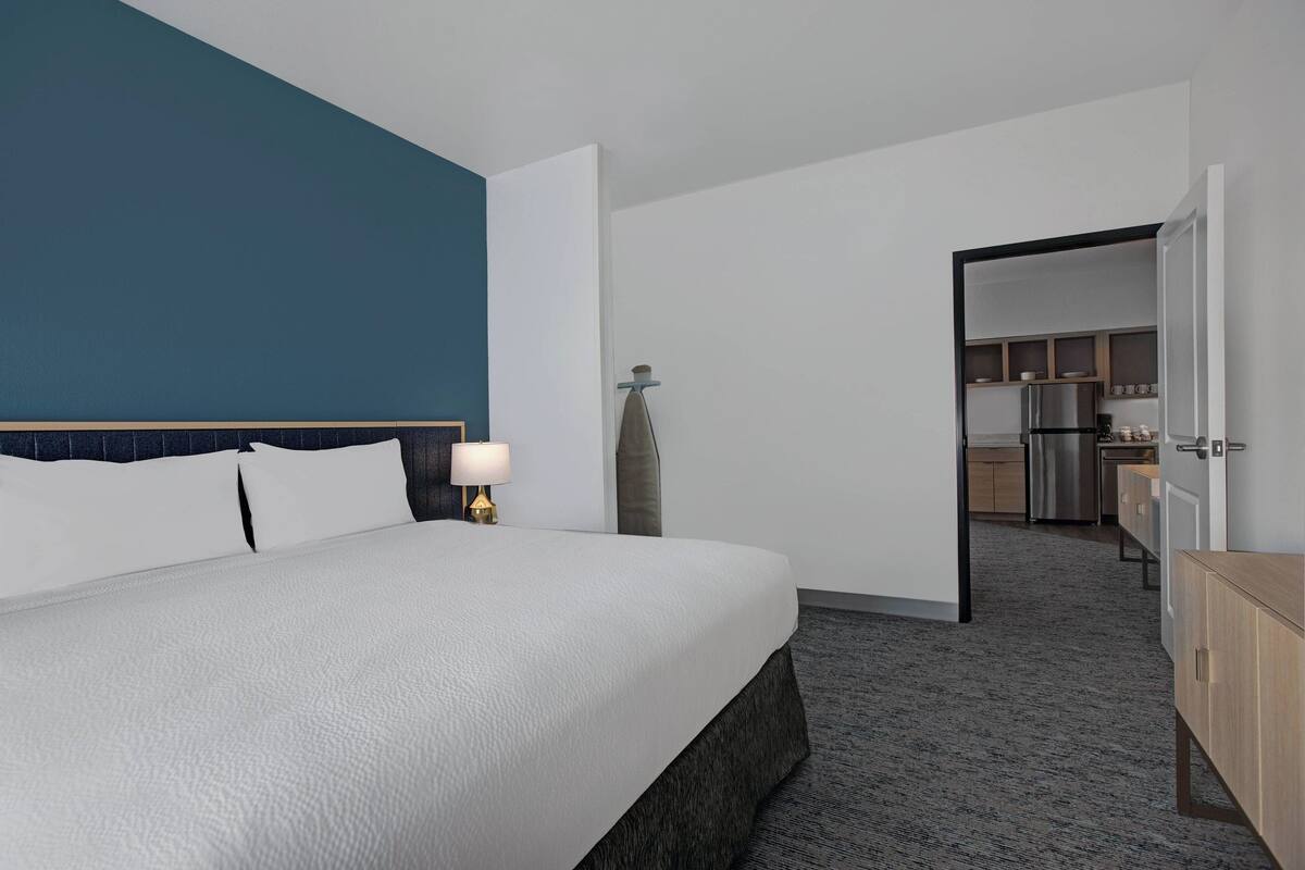 Habitación de hotel con una cama grande, una mesita de noche con lámpara, un armario y una kitchenette al fondo.