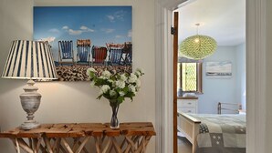 A room with a wooden console table, a lamp, a vase with flowers, a beach-themed painting, a bed with a star-patterned blanket, and a hanging light fixture.