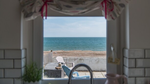 Blick aus einem Fenster auf einen Strand mit einer Holzterrasse und Stühlen.